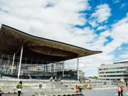 Ffotograff o du blaen adeilad y Senedd ym Mae Caerdydd. Mae'r adeilad wedi'i wneud o frics llwyd, ac mae rhai trawstiau gwyn i'w gweld. Mae arwydd sy'n dweud 'Senedd Cymru'. Yn y blaendir, mae cerddwyr a beicwyr yn mynd heibio. Mae rhai pobl yn eistedd ar y grisiau y tu allan i adeilad y Senedd.