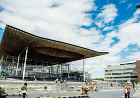 Ffotograff o du blaen adeilad y Senedd ym Mae Caerdydd. Mae'r adeilad wedi'i wneud o frics llwyd, ac mae rhai trawstiau gwyn i'w gweld. Mae arwydd sy'n dweud 'Senedd Cymru'. Yn y blaendir, mae cerddwyr a beicwyr yn mynd heibio. Mae rhai pobl yn eistedd ar y grisiau y tu allan i adeilad y Senedd.